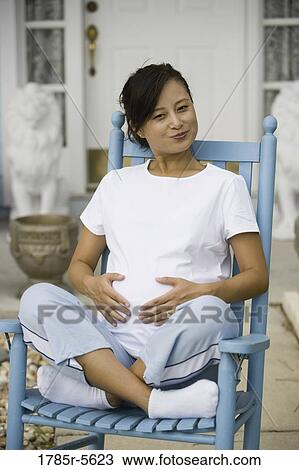 Stock Image - Pregnant woman sitting on a rocking chair in front of house. Fotosearch
