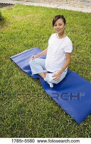 Stock Image - Pregnant woman sitting on exercise mat on grass. Fotosearch