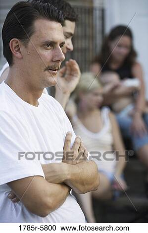 Stock Image - Profile of contemplative man with folded arms with family and friends in background. Fotosearch