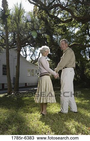 Stock Image - Rear view of mature couple holding hands under oak trees. Fotosearch
