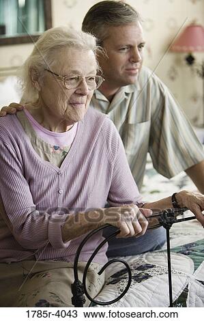 Stock Image - Senior woman and adult son sitting on bed at nursing home. Fotosearch