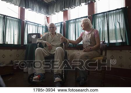 Picture - Senior woman sitting next to feeble senior man in wheelchair in bedroom. Fotosearch