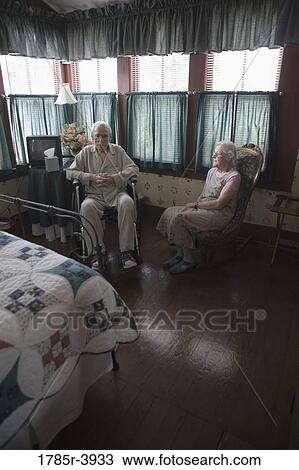 Stock Image - Senior woman sitting next to feeble senior man in wheelchair in bedroom. Fotosearch