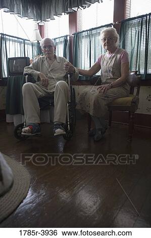 Stock Photograph - Senior woman sitting next to feeble senior man in wheelchair in bedroom. Fotosearch