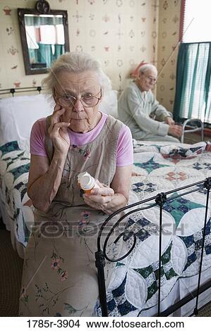 Picture - Senior woman sitting on bed looking at prescription bottle. Fotosearch