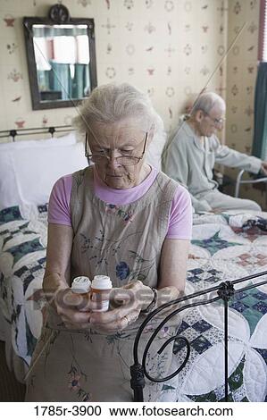 Stock Image - Senior woman sitting on bed looking at prescription bottle. Fotosearch