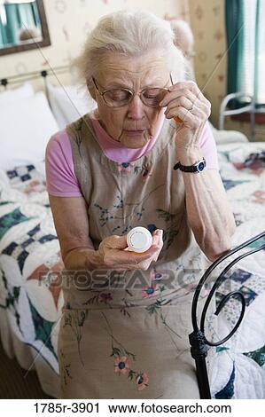 Stock Image - Senior woman sitting on bed looking at prescription bottle. Fotosearch