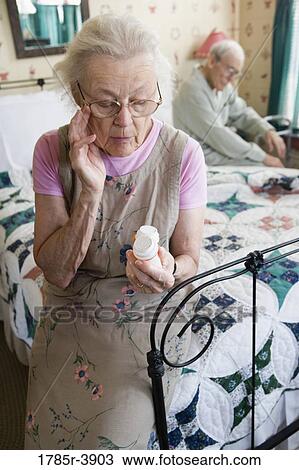 Stock Image - Senior woman sitting on bed looking at prescription bottle. Fotosearch