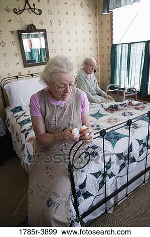 Stock Photo - Senior woman sitting on bed looking at prescription bottle. Fotosearch