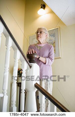 Stock Photo - Senior woman standing by staircase alone, looking up the stairs. Fotosearch