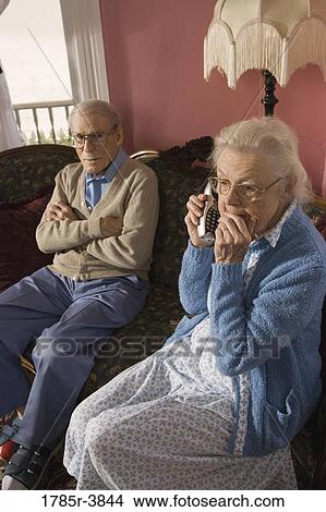 Picture - Senior woman talks on cordless phone while grumpy senior man watches. Fotosearch