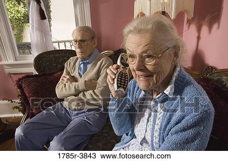 Stock Image - Senior woman talks on cordless phone while grumpy senior man watches. Fotosearch