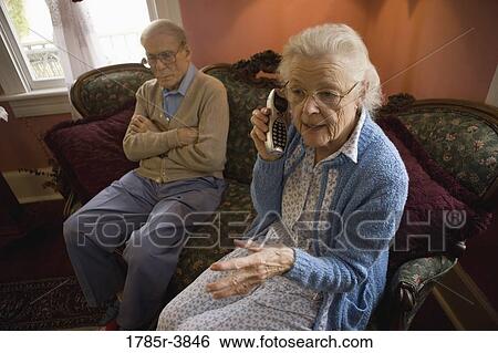 Stock Photograph - Senior woman talks on cordless phone while grumpy senior man watches. Fotosearch