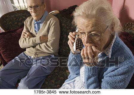 Stock Photography - Senior woman talks on cordless phone while grumpy senior man watches. Fotosearch