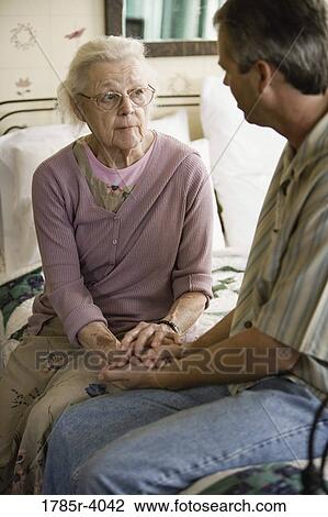 Stock Image - Senior woman with adult son conversing while sitting on bed. Fotosearch