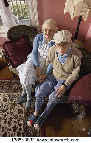 Stock Image - Smiling senior couple sitting on sofa in living room. Fotosearch