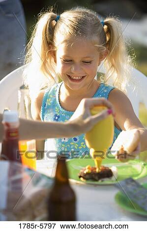 Smiling young girl looking on as somebody puts mustard on her burger View Large Photo Image Stock Photo - Smiling young girl looking on as somebody puts mustard on her burger. Fotosearch