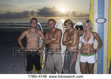 Stock Photograph - Surfer dudes and girl posing on beach at sunrise with surfboards. Fotosearch