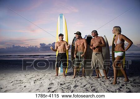 Stock Photography - Surfer dudes and girl posing on beach at sunrise with surfboards. Fotosearch