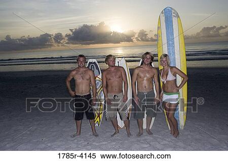 Stock Photography - Surfer dudes and girl posing on beach at sunrise with surfboards. Fotosearch