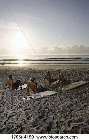 Stock Image - Surfer dudes and girl sitting on beach at sunrise with surfboards. Fotosearch