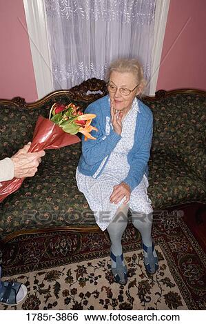 Stock Photograph - Surprised and happy senior woman sitting on the sofa handed bouquet of flowers. Fotosearch