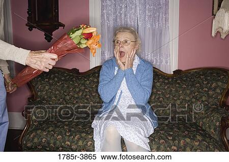 Stock Photography - Surprised and happy senior woman sitting on the sofa handed bouquet of flowers. Fotosearch