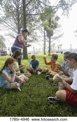 Stock Photograph - Teacher distributing cupcakes to children seated in a circle at a park. Fotosearch