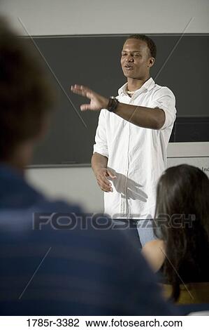 Teacher standing in the classroom gesturing towards class View Large Photo Image Stock Image - Teacher standing in the classroom gesturing towards class. Fotosearch