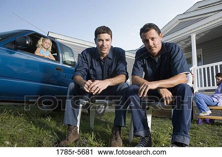 Stock Image - Two blue-collar men holding cold drinks sitting on lawn chairs in front of trailer home. Fotosearch