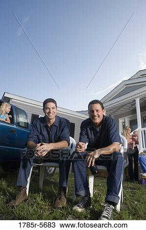 Stock Image - Two blue-collar men holding cold drinks sitting on lawn chairs in front of trailer home. Fotosearch