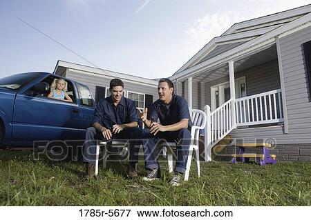 Stock Photo - Two blue-collar men holding cold drinks sitting on lawn chairs in front of trailer home. Fotosearch