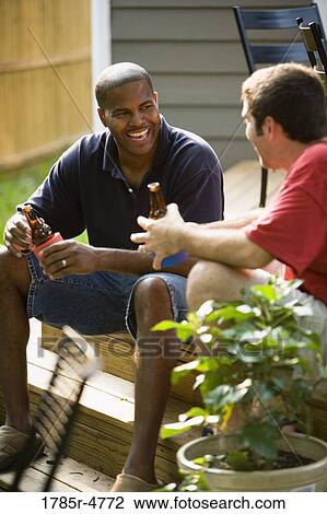 Stock Image - Two men sitting on steps of backyard patio enjoying cold beverages. Fotosearch
