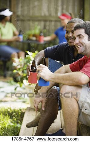 Stock Photo - Two men sitting on steps of backyard patio enjoying cold beverages. Fotosearch