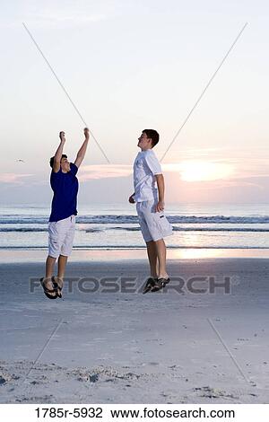 Stock Image - Two teenage boys playing and jumping on the beach at sunrise. Fotosearch