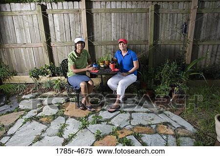 Stock Photography - Two women sitting together around backyard patio table with cold beverages. Fotosearch