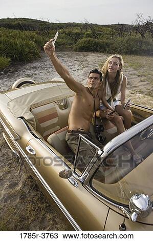 Stock Image - View of a young couple in antique convertible car looking at photographs. Fotosearch