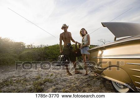 Stock Image - View of a young couple standing by antique convertible car. Fotosearch