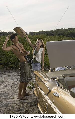 Stock Photograph - View of a young couple standing by antique convertible car. Fotosearch