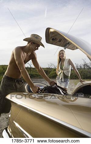 Stock Image - View of a young couple unloading bags from trunk of antique car. Fotosearch