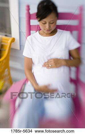 Picture - View of pregnant woman sitting in rocking chair looking at her belly. Fotosearch