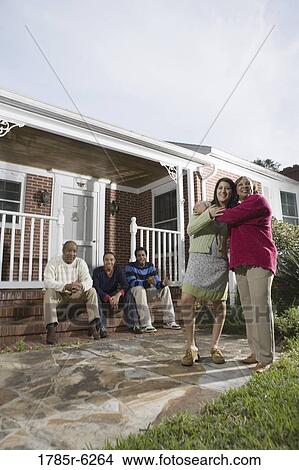 Picture - Woman embracing her daughter-in-law in front of house with family in background. Fotosearch