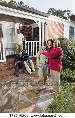 Stock Image - Woman embracing her daughter-in-law in front of house with family in background. Fotosearch