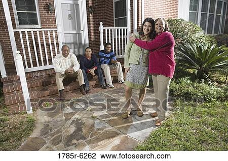 Stock Image - Woman embracing her daughter-in-law in front of house with family in background. Fotosearch