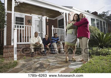 Woman embracing her daughter-in-law in front of house with family in background View Large Photo Image Stock Image - Woman embracing her daughter-in-law in front of house with family in background. Fotosearch