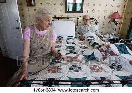 Picture - Worried senior woman sitting on bed with bedridden senior man. Fotosearch
