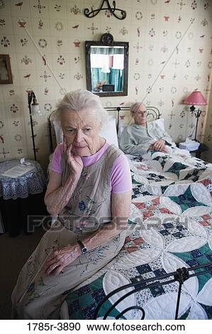 Stock Image - Worried senior woman sitting on bed with bedridden senior man. Fotosearch