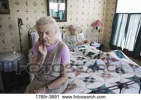 Stock Image - Worried senior woman sitting on bed with bedridden senior man. Fotosearch