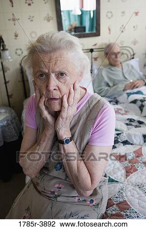 Stock Image - Worried senior woman sitting on bed with bedridden senior man. Fotosearch
