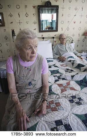 Stock Image - Worried senior woman sitting on bed with bedridden senior man. Fotosearch